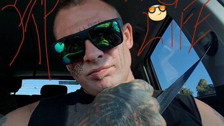 A heavily tattooed man sitting in a car, taking a selfie.