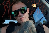 A heavily tattooed man sitting in a car, taking a selfie.