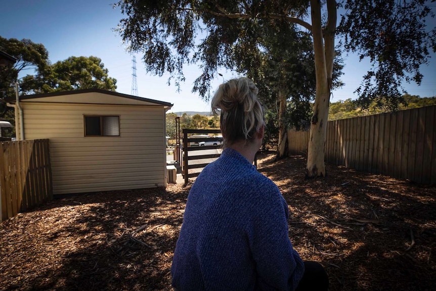 A woman sits on a bench with her back facing the camera with a caravan park cabin and trees in the background.