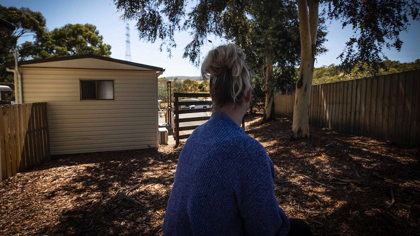 A woman sits on a bench with her back facing the camera with a caravan park cabin and trees in the background.