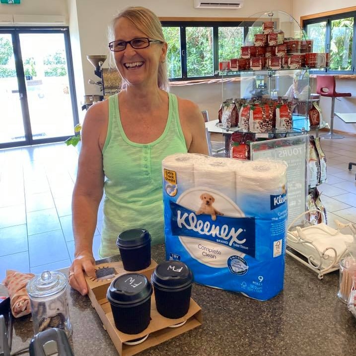 Smiling woman stands at a cafe counter with a nine-pack of Kleenex toilet paper and three takeaway coffees.