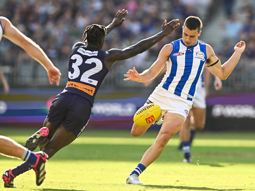 A North Melbourne midfielder looks down at the ball as he goes through a kick as a defender reaches out to smother.