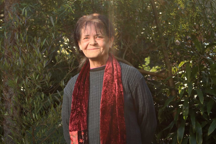 A woman with dark hair and a long red scarf stands amongst the trees, smiling.