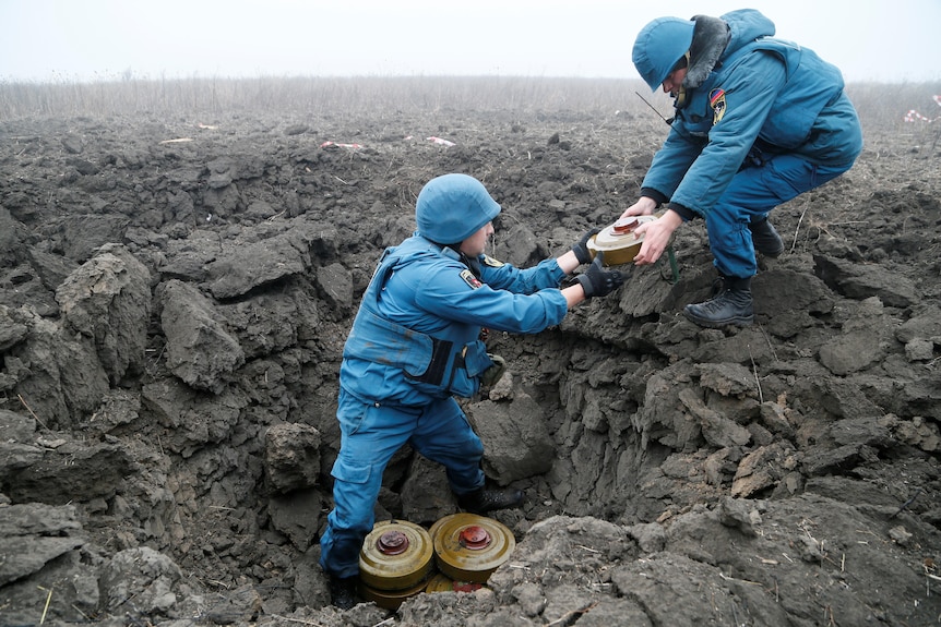 Two emergency workers pass anti-tank mines to each other from a crater in a desolate, muddy field. 