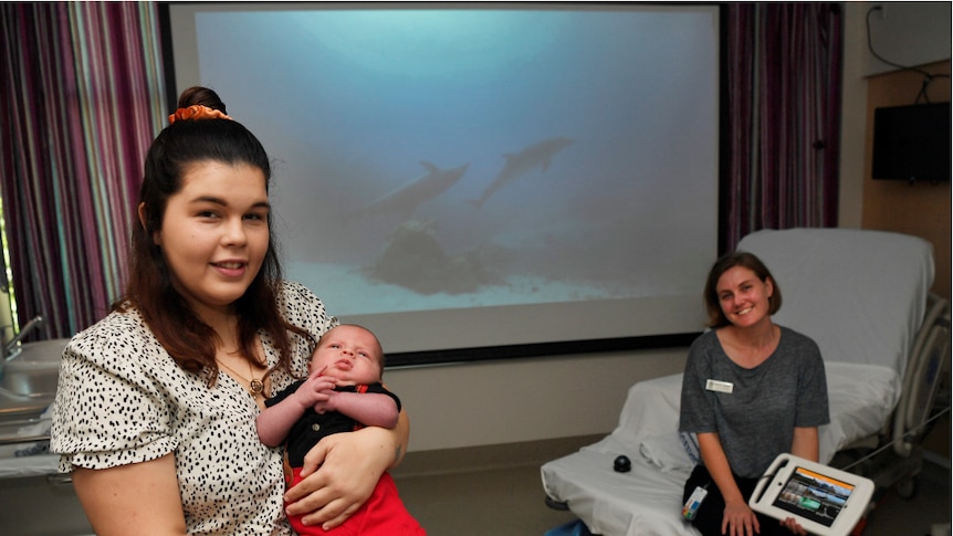 Brunette woman holding baby in hospital birthing room next to blonde midwife in front of a projector screen showing dolphins.