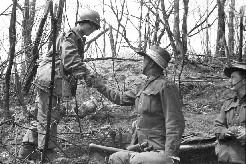 Two soldiers shake hands, wearing uniforms in bushland, in a black and white photo.