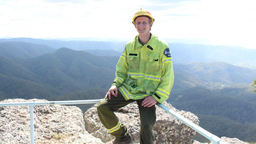 A young man, wearing a helmet and high viz jacket, sits in front of a view will hills