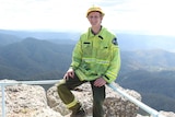 A young man, wearing a helmet and high viz jacket, sits in front of a view will hills