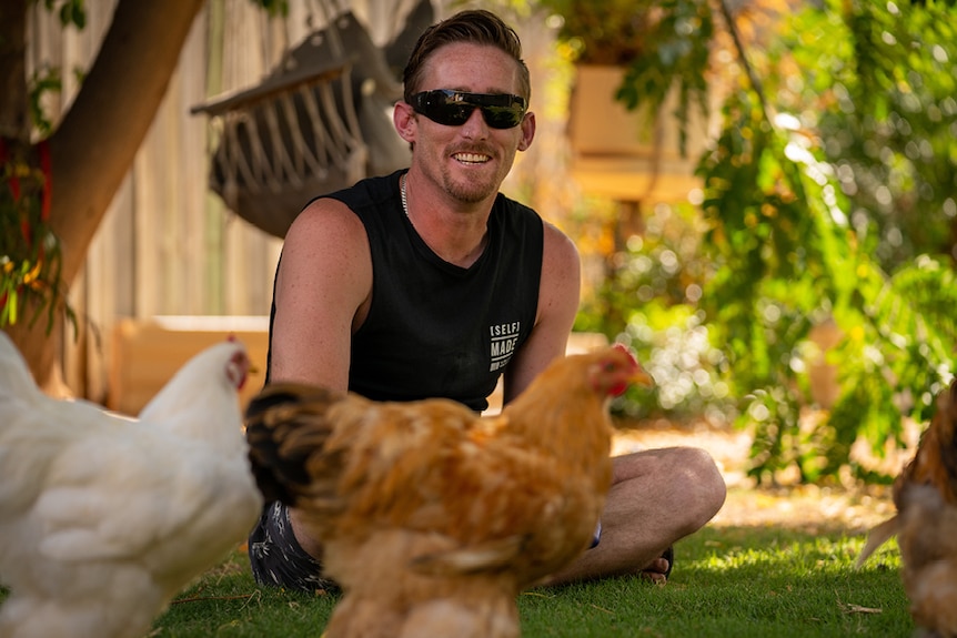 A man wearing sunnies sitting with three chickens in a yard