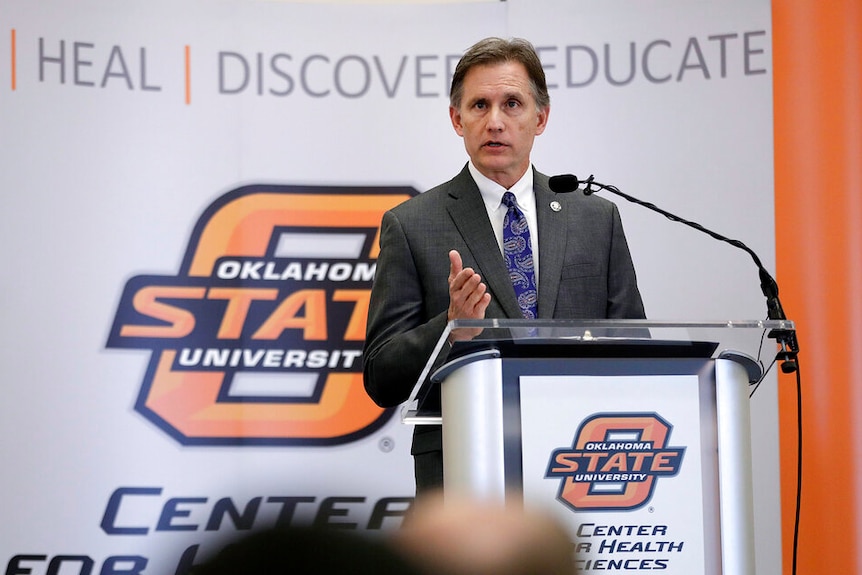 A man in a suit stands at a lectern in front of Oklahoma State University branding.
