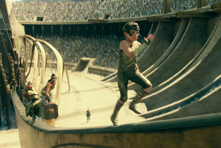 Little girl in gladiator-style costume runs up ramp with massive arena, full of people, in background behind her.