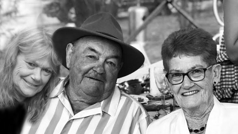A woman leans against an older man in a hat and striped shirt and an older woman wearing glasses, all smiling.