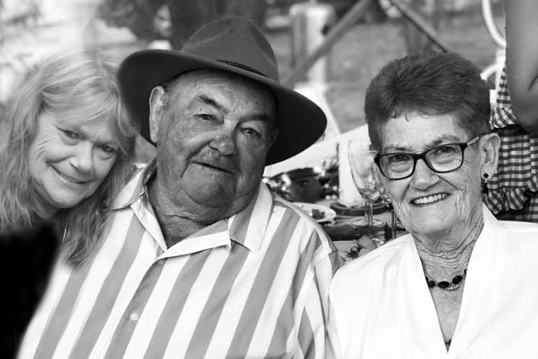 A woman leans against an older man in a hat and striped shirt and an older woman wearing glasses, all smiling.