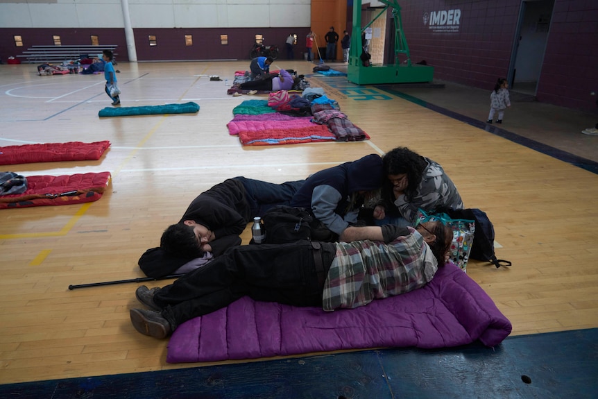 People lie on sleeping bags that are on a gymnasium floor.