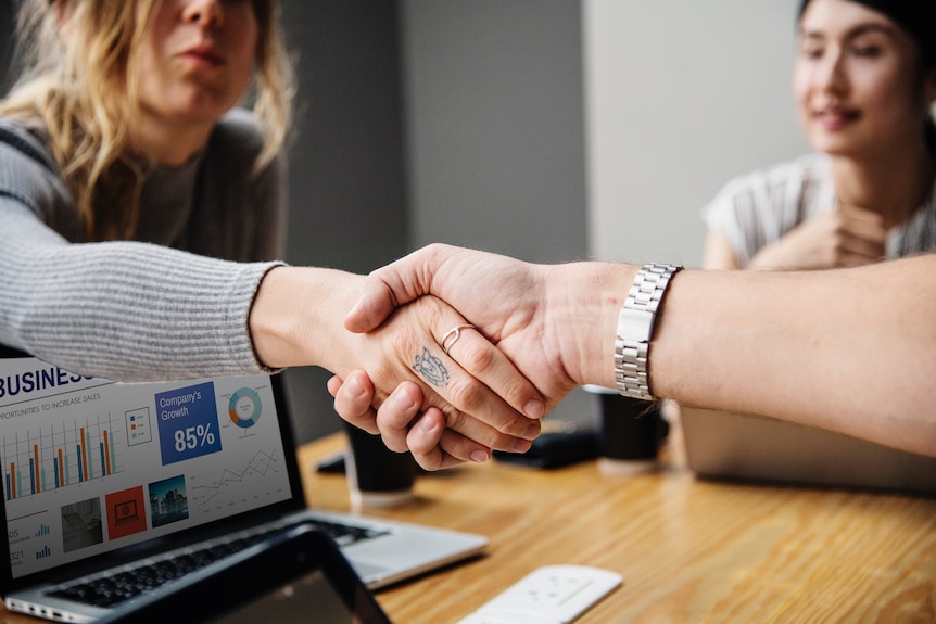 Two women shaking hands across a table. One woman has a small tattoo on her finger.