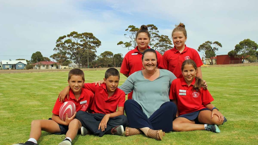 Five school students in red uniforms sit on new green grass with one of their female teachers.