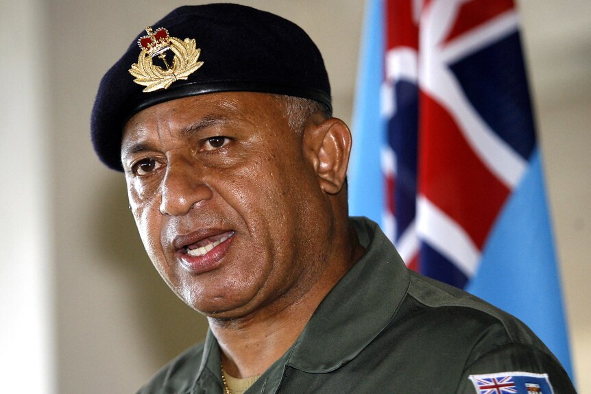 A man in a green military shirt and a black beret, with the Fijian flag behind him.