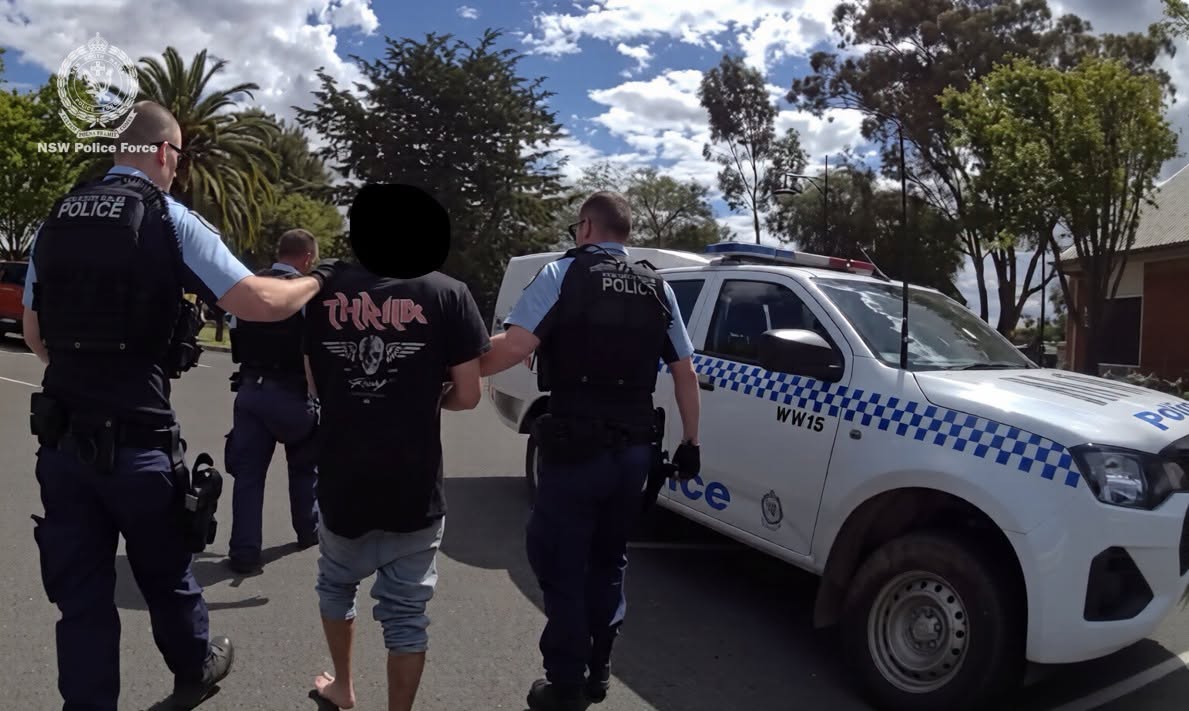 Three men walking with their backs turned, two wearing police vests, the man in the middle wearing a black shirt.