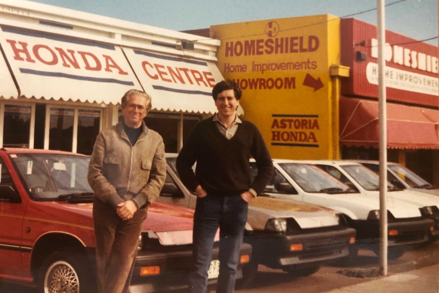 Two men stand in front of early-1980s Honda cars at a car dealership.