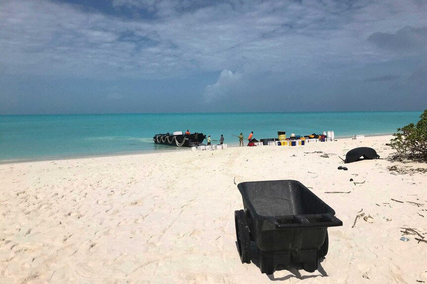 Bright blue water is seen under cloudy blue skies with light sand on the beach in front. A crew is seen on a boat and on shore.