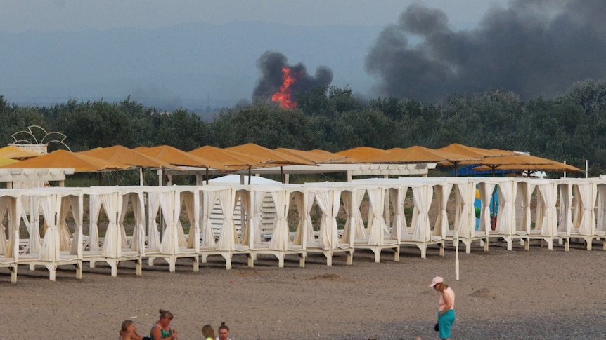 People rest on a beach as smoke and flames rise after an explosion in the background.