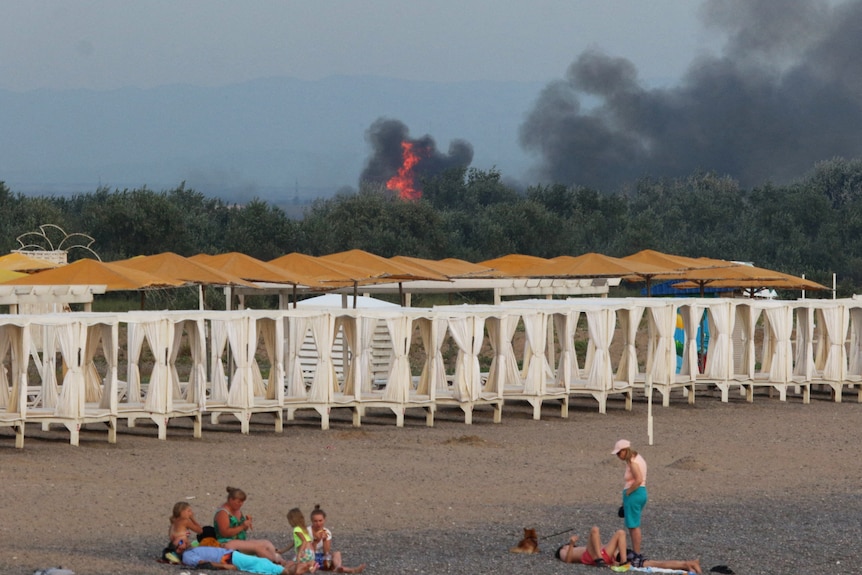 People rest on a beach as smoke and flames rise after an explosion in the background.