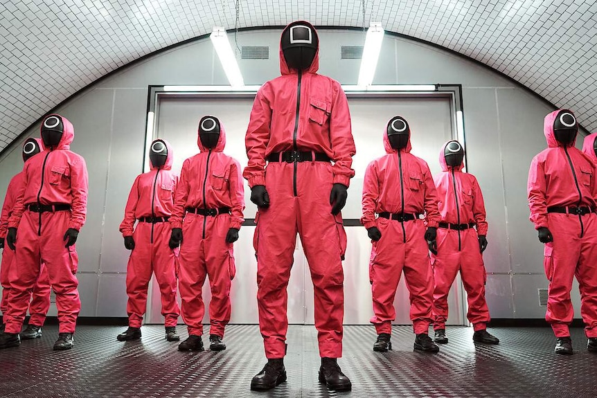 A film still from Netflix series Squid Game, depicting 9 figures in pink uniforms wearing black masks with circles and squares.