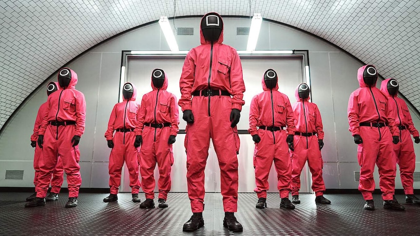 A film still from Netflix series Squid Game, depicting 9 figures in pink uniforms wearing black masks with circles and squares.