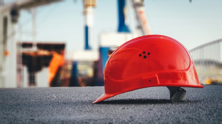 An orange construction hat sits on concrete with an out of focus construction site in the background.