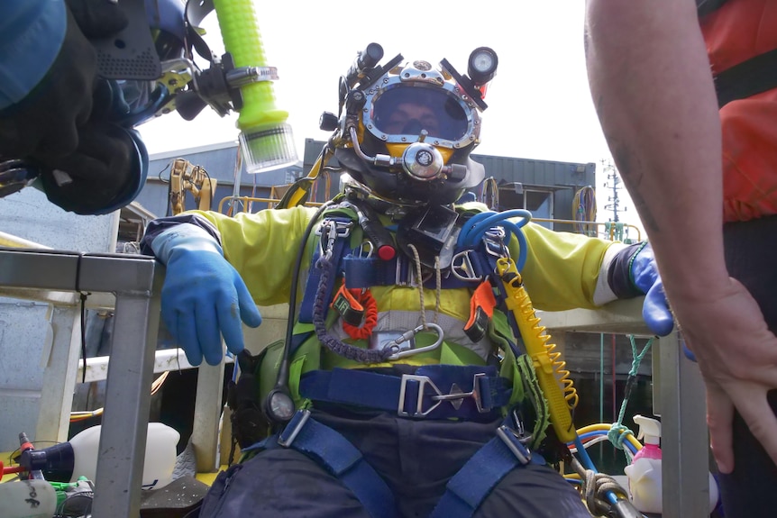A person sitting in a very cumbersome-looking commercial diving suit, with helmet.
