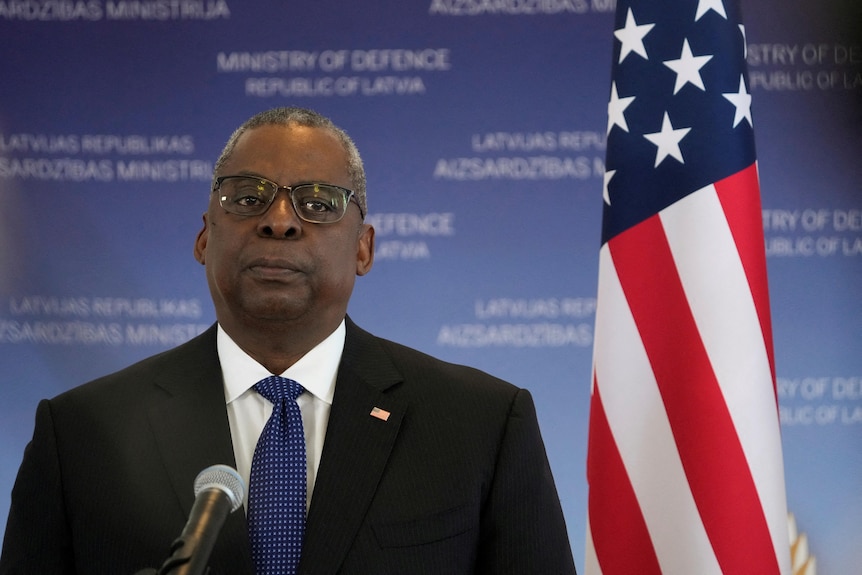 A sombre-looking man standing behind a microphone, next to a USA flag.