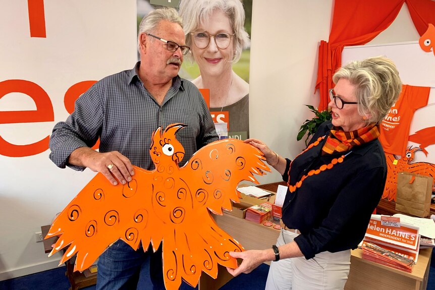 A man holds up an orange cardboard cut-out of a cockatoo, while a independent candidate for Indi Helen Haines inspects it.