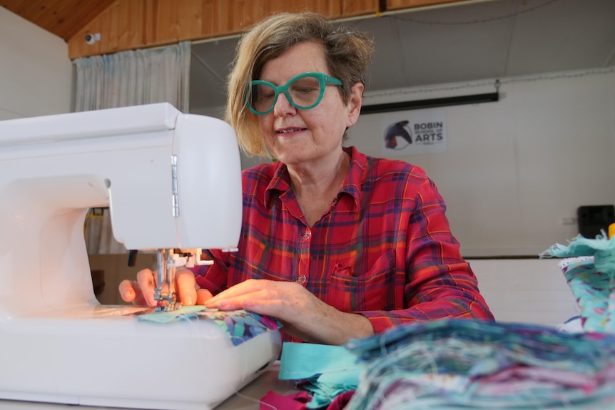 A woman sits sewing, with piles of material near her.