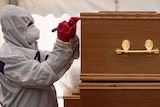 A person in a HAZMAT suit and mask marks a coffin in a tent.