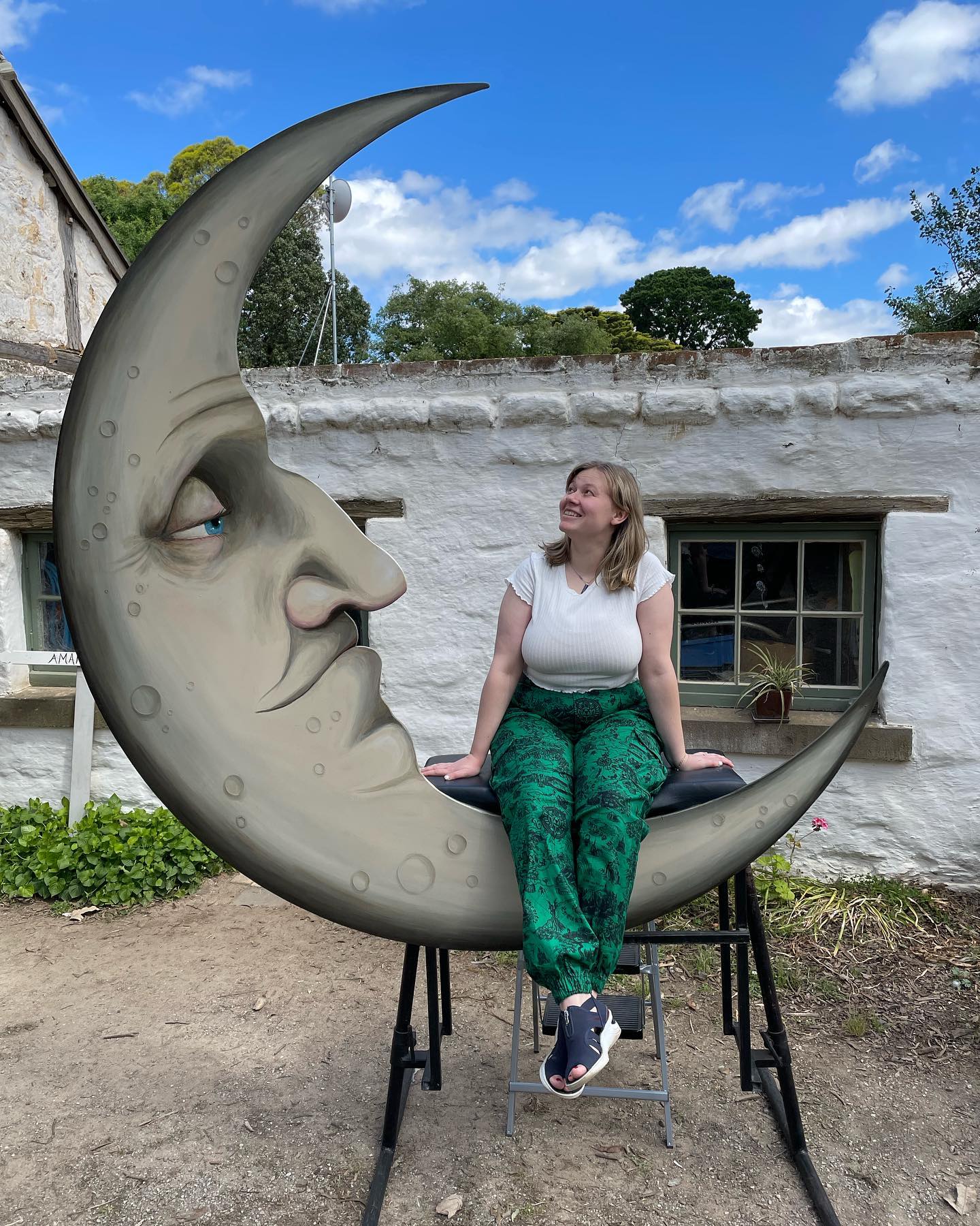 Silvi Vann-Wall sits on a large wooden cutout of a moon with their legs crossed, smiling. 