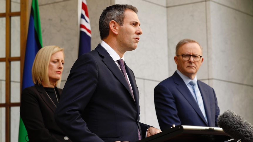 Two men and a woman in suits stand behind podiums in front of a doorway into Parliament House.
