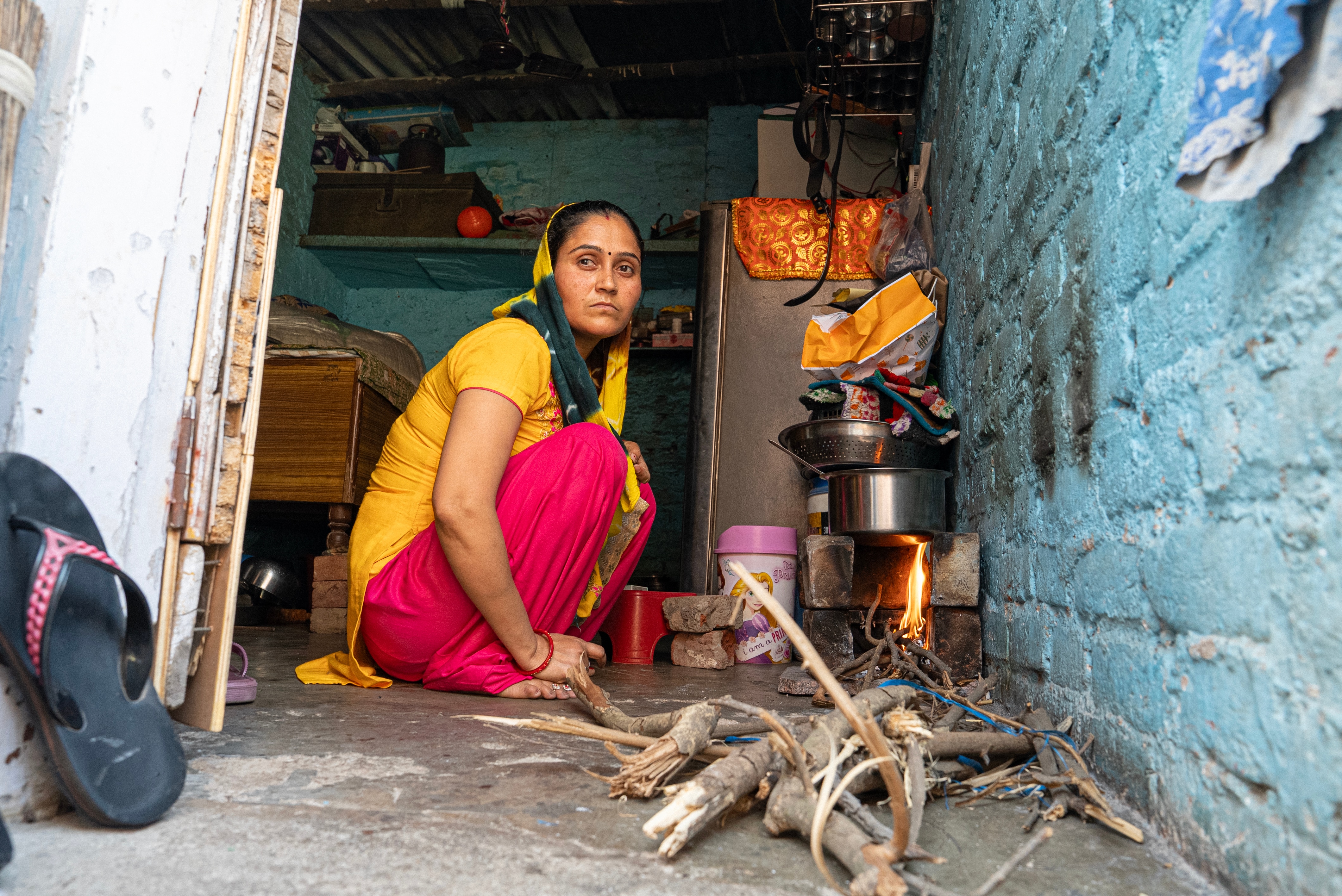 Serious Pooja dressed in yellow sits over a floor with stick next to a wood burning stove, utensil on top. 