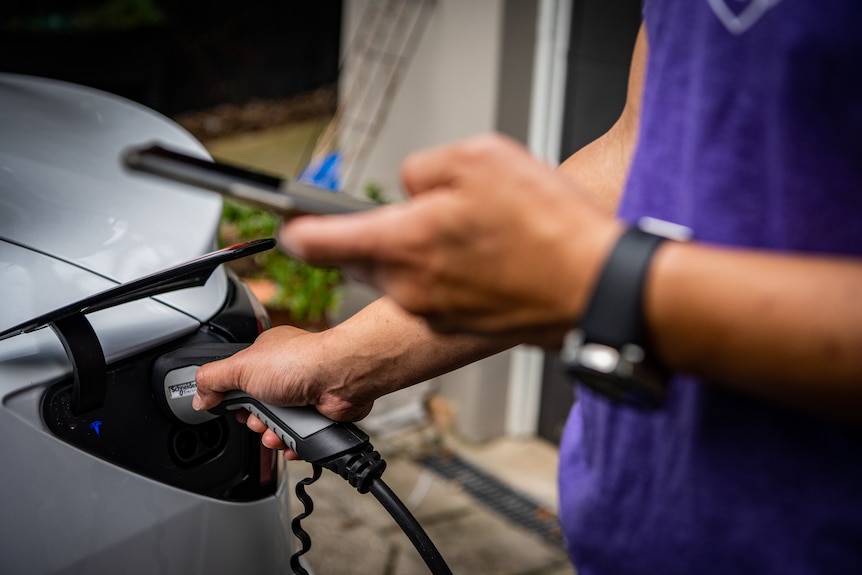 A close-up of a man checking the charging status of his car on his phone while plugging a charger into the car.