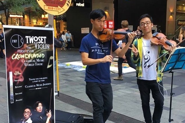 Brett and Eddy busking in a mall, TwoSet Violin crowdfunded sign next to them.