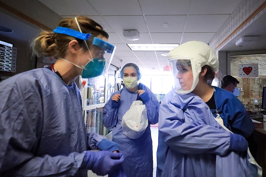 medical staff in masks and gowns and PPE look at each other in a hospital ward with sun coming in from a window