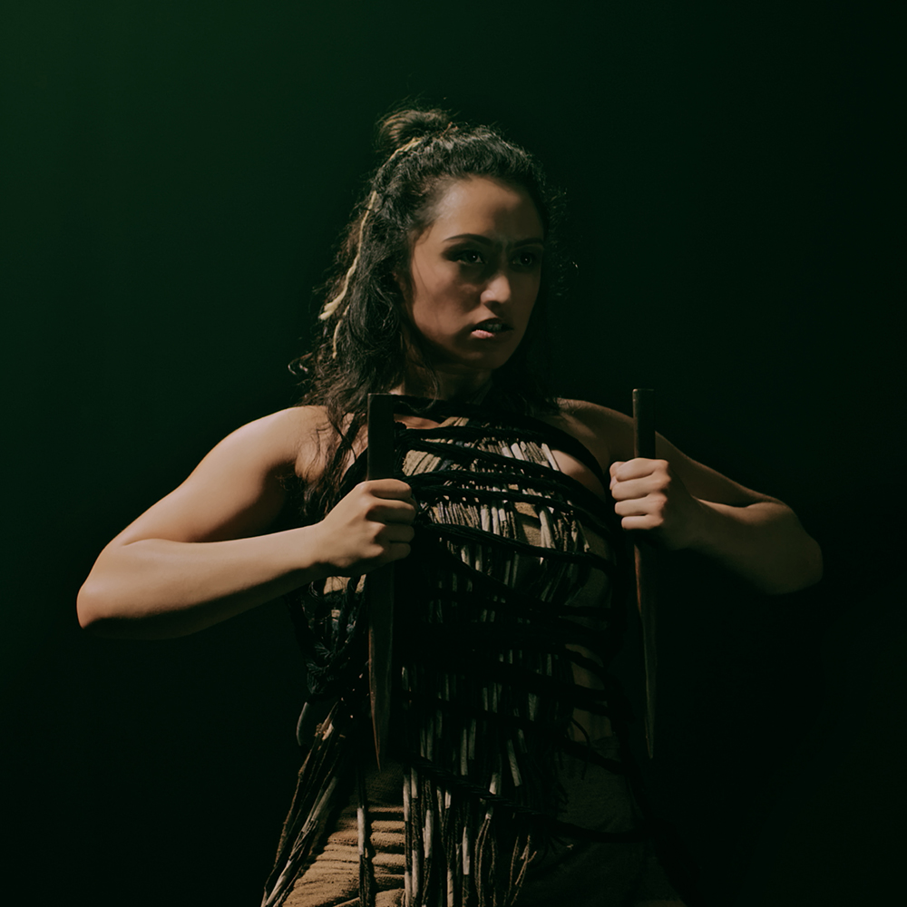 A woman with half hair up, half in bun, wears traditional Māori attire and holds two staff weapons in each hand close to chest.