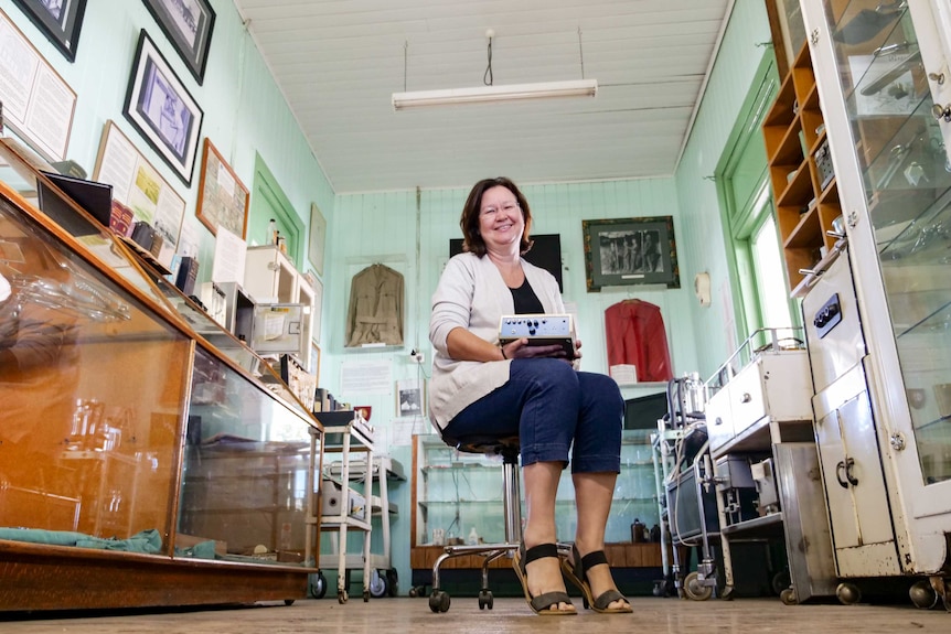 A woman sitting on a stool holds a piece of medical equipment, sitting in an old museum.