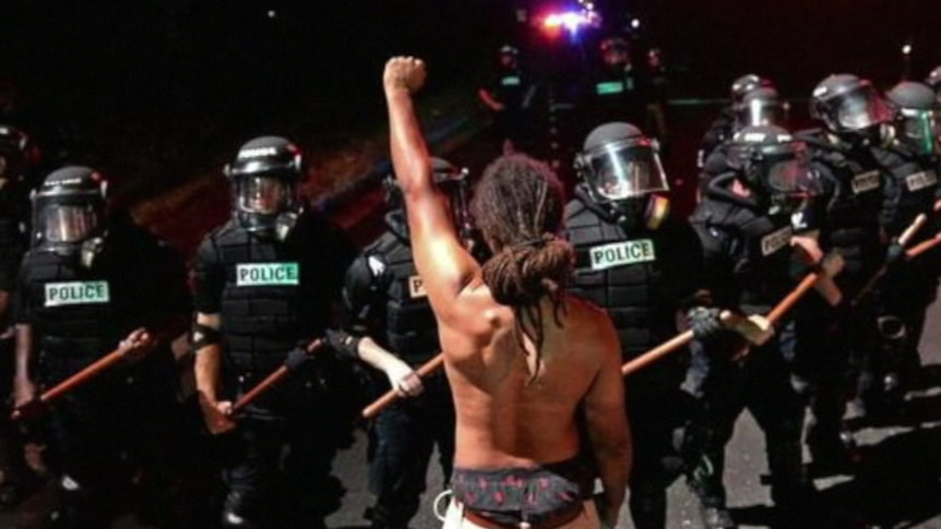 A black man stands in front of a line of riot police, shirtless, with his fist raised.