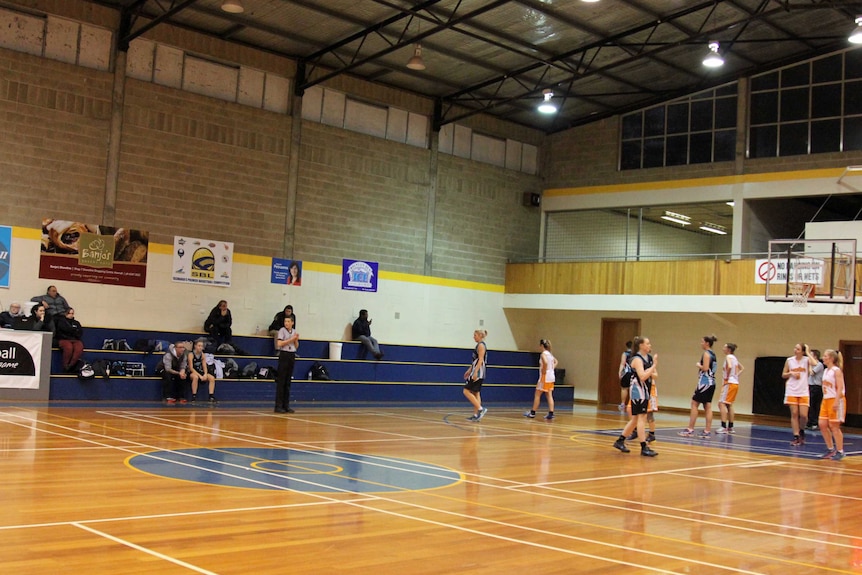 A wide shot of a suburban gymnasium basketball court, players standing under the ring, with a few crowd members in the stands.