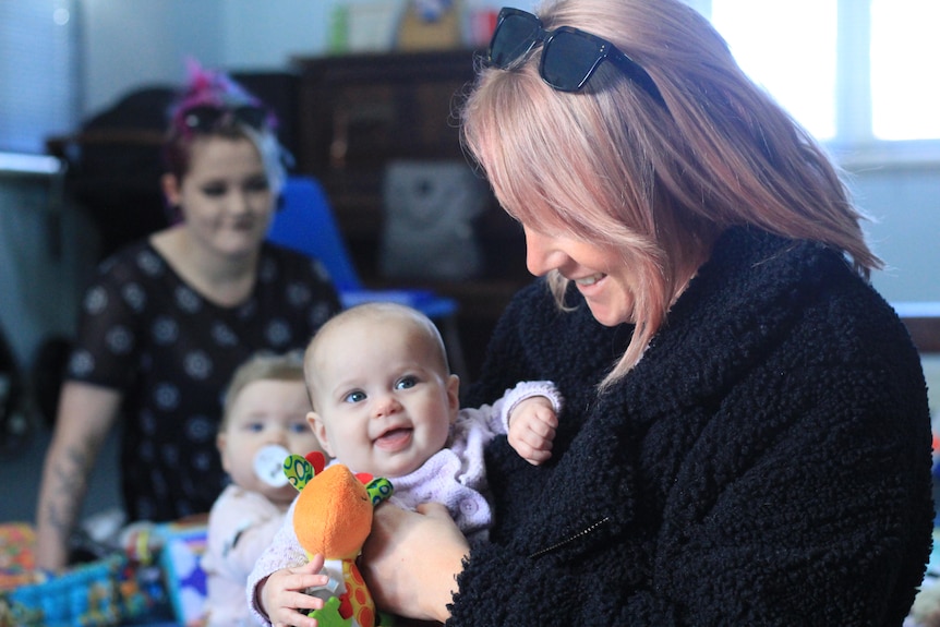 A lady with pink hair is holding a baby, in the background there's a younger woman and a baby, sitting on the floor.