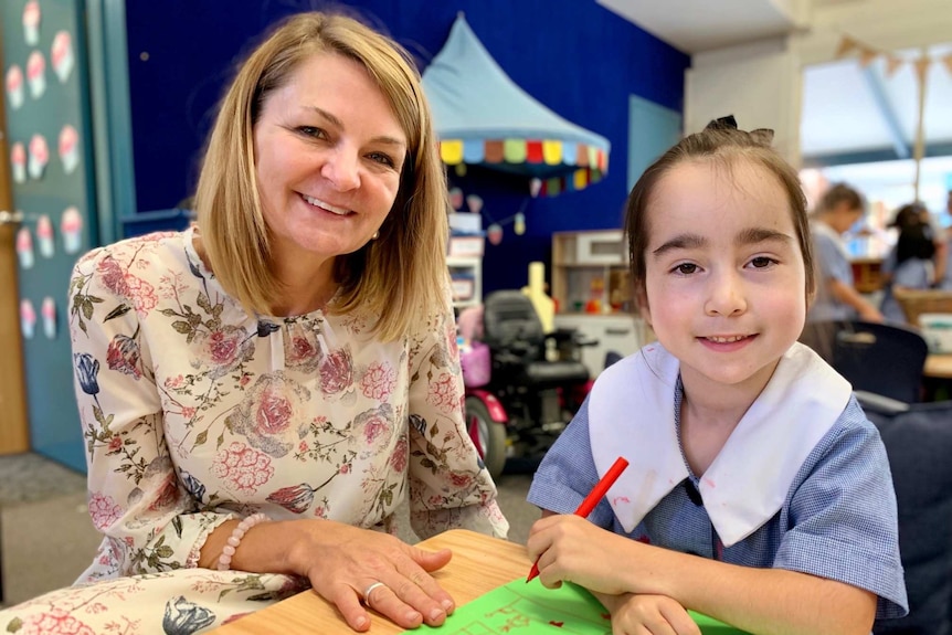 A young girl in a school uniform sits at a table next to female teacher. The girl has a crayon in her hand.