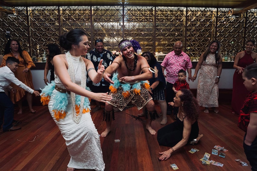 A woman dances next to a man wearing traditional Samoan outfit surrounded by a group of people pictured laughing.