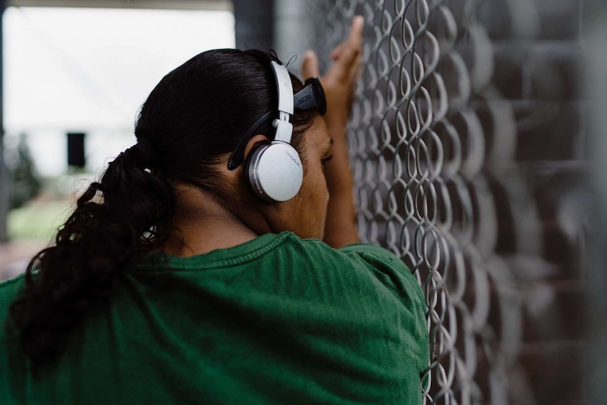 A woman wears headphones while looking out through the chain link fence in prison.