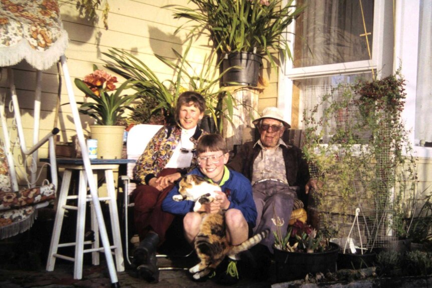 An older man and woman sit in a leafy backyard with a young boy, who is holding a cat.