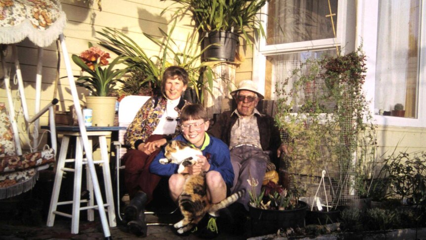 An older man and woman sit in a leafy backyard with a young boy, who is holding a cat.